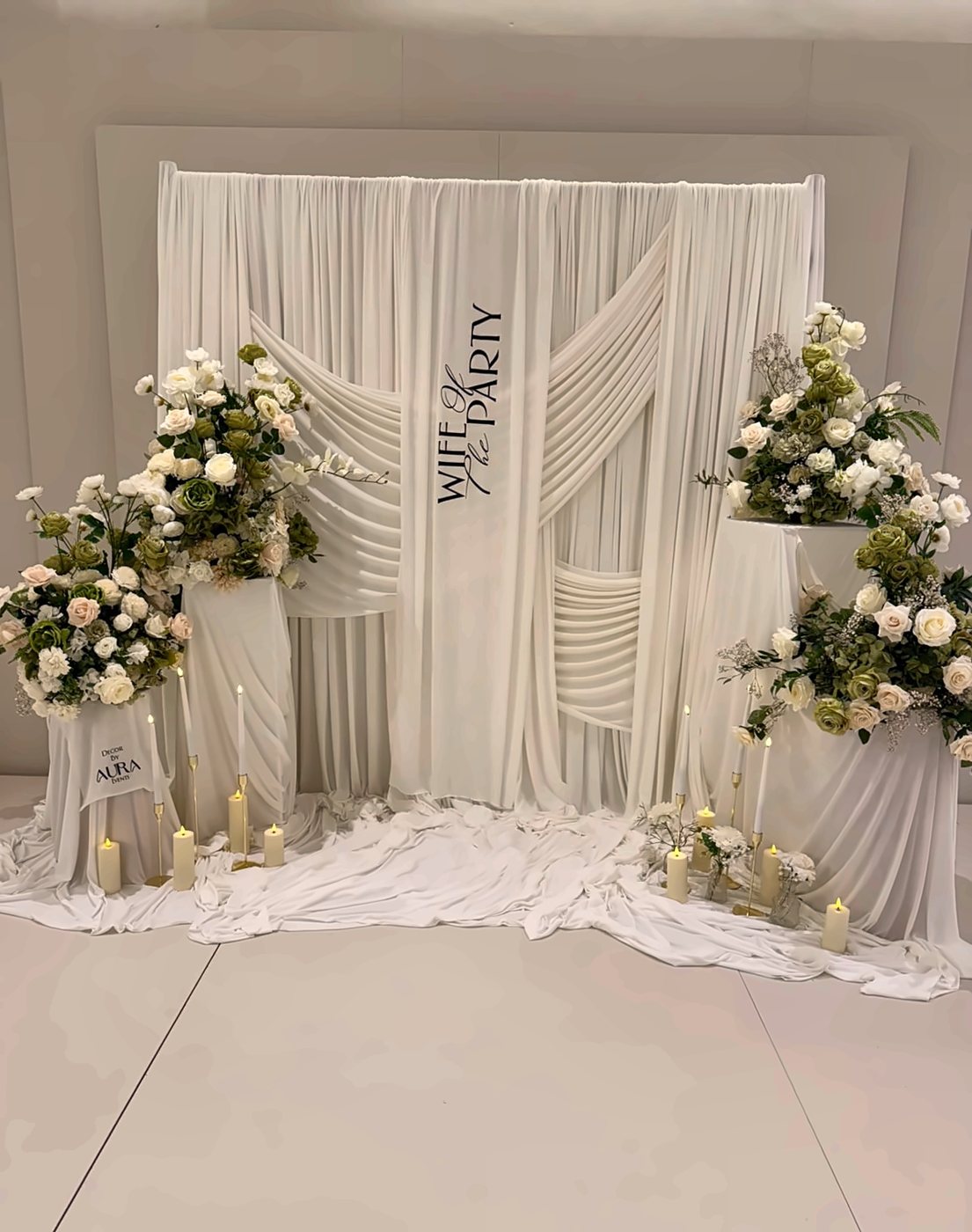 A white draped backdrop with florals and candles, styled for a bridal shower at The XL Suite