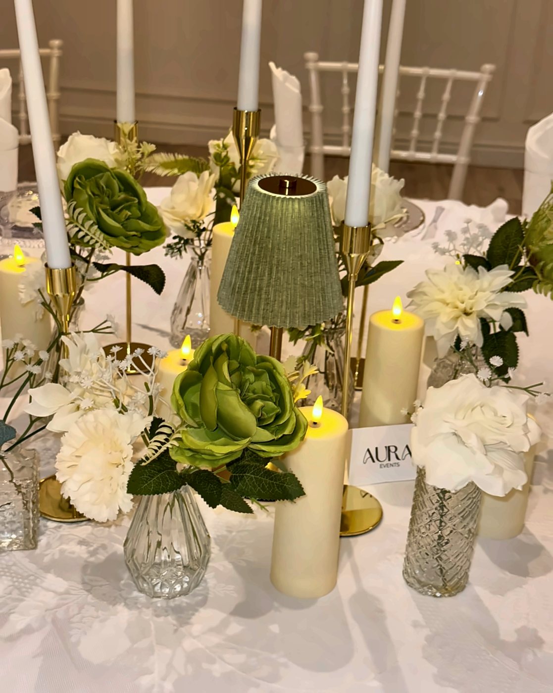 A table centrepiece of green and white roses, candles and a small lamp