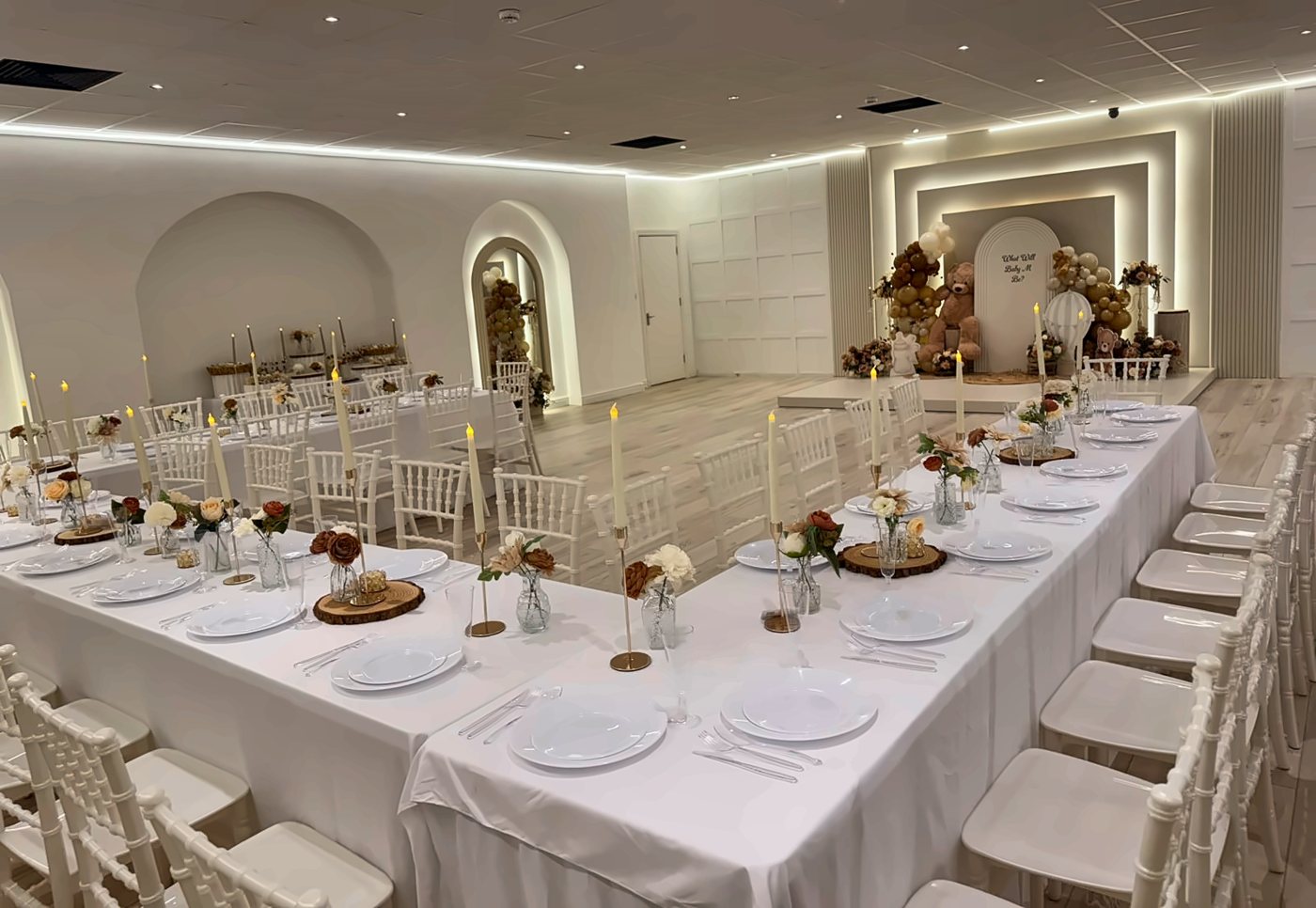 Long tables in a U with candles and rust-and-cream florals, a teddy-bear arch on the stage, set for a gender reveal