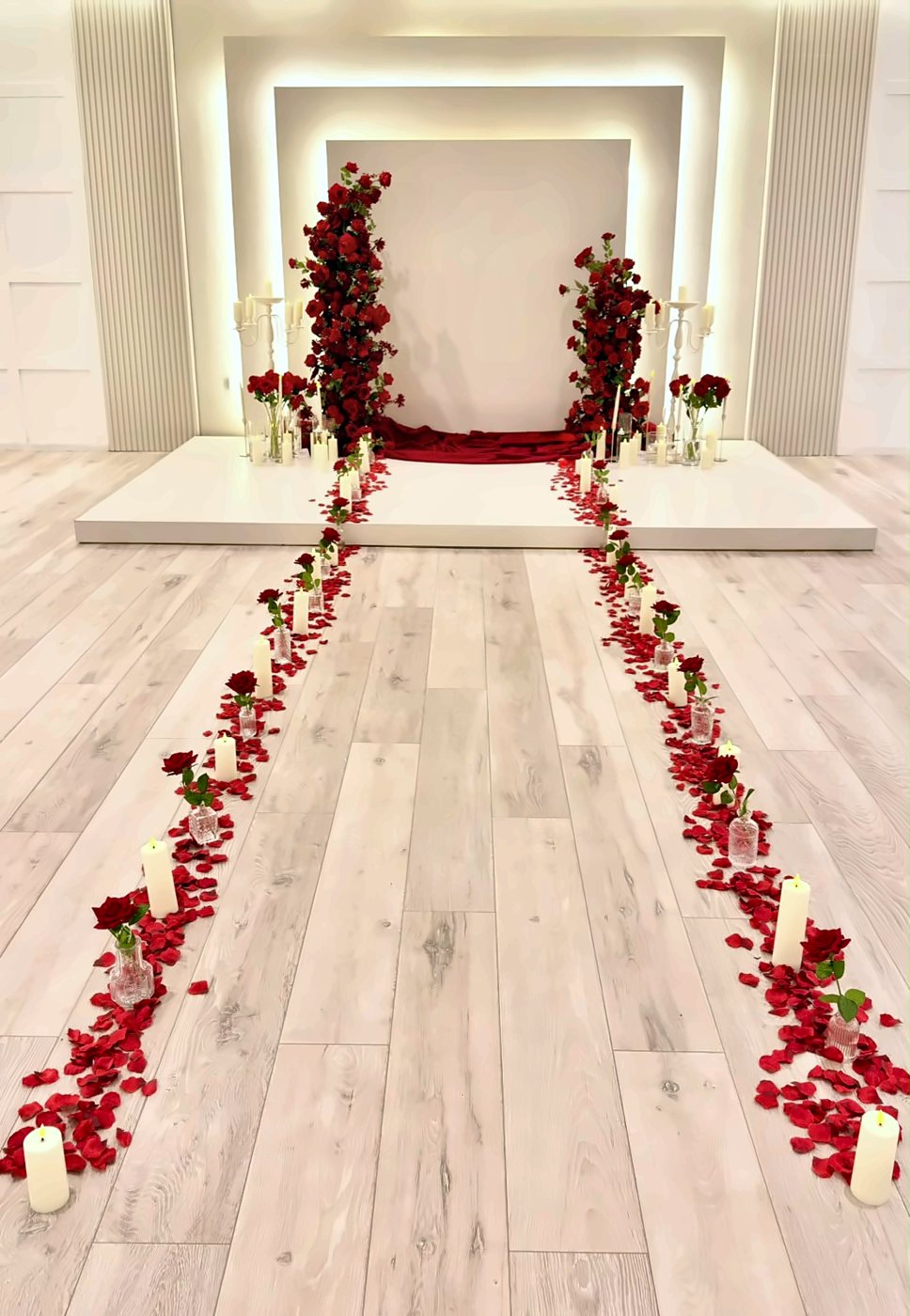 A candlelit aisle of red rose petals leading to a white stage dressed with red roses, set for a proposal